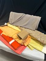 Seven bolts of fabric in a pile, showing colors and patterns including beige, yellow, orange, and a geometric pattern.