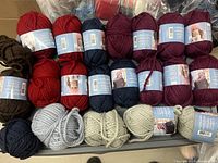Photo showing multiple balls of knitting yarn in colors red, purple, navy blue, brown, gray, cream, and maroon arranged in rows.