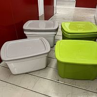 Four beige plastic storage totes with lids stacked and four green plastic storage totes stacked next to them on floor.