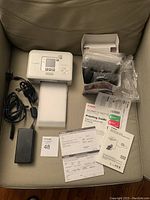 Canon Selphy CP photo printer on a beige chair with power adapter, USB cable, new ink cartridge and paper, and printed manuals arranged around it.