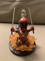 Front view of Deadpool statue holding two swords, mounted on translucent orange smoke/explosive effect base