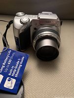 Front view of the Olympus silver and black digital camera with zoom lens extended, visible tags indicating battery life and shooting capacity.