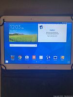 Front view of Samsung Galaxy Tab 4 tablet turned on showing the home screen with app icons and time/date as 12:03 PM, May 7.