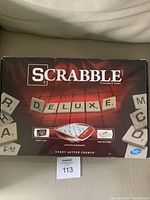 Front of Scrabble Deluxe board game box showing the word DELUXE formed with tiles and highlighting the carry case, rotating gameboard, and non-slip grid features