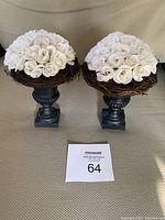 Two identical flower pedestal arrangements with white artificial roses atop black pedestals resembling urns, placed side by side on a neutral fabric surface with a lot number card in front.