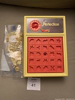 Complete vintage Perfection game with beige base, red shape board and timer dial, along with bag of yellow shape pieces.