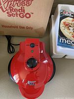 Closed view of red Xpress Redi Set Go electric grill cooker showing control knob with timer and power switch with indicator light, beside original box and instruction manual.