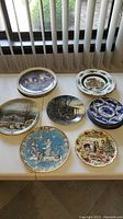 Sixteen collector plates displayed on table showing detailed artwork and various themes, including Christmas and Americana scenes.