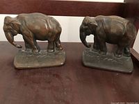Photo showing two identical heavy metal elephant bookends side by side on a dark wooden surface.