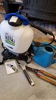 Photo showing Field King backpack sprayer, blue plastic watering can, garden shears, stapler, Dewalt utility knife, water testing kit documents, and blue winter gloves on top of sprayer.