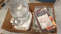 Photo showing the Kitchen Aid food processor with large clear plastic bowl and feed chute, instruction and recipe booklet, smaller bowl, and a box of attachments.