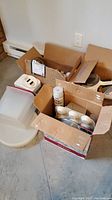 Photo showing cardboard boxes filled with metal baking tins, cookie cutters, plastic ashtrays, spatula, and various kitchen gadgets on floor