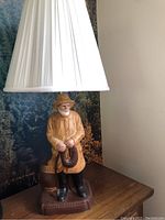 Full lamp showing the white pleated lampshade and the figurine base depicting a bearded man in a yellow coat and hat, holding a coiled rope while standing.