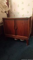 Closed wooden nightstand with two doors and curved handles, glass top visible. Warm brown tone with some wear.