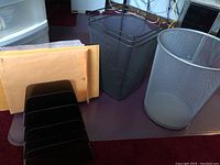 Two black mesh metal trash cans, manila envelopes, and black acrylic desk organizer on a purple surface