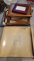 Stack of twelve wooden picture frames with a smaller red wooden frame on top, and a square glass serving dish with engraved 'H' in the foreground.