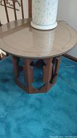 Side table full view showing round glass top and hexagonal carved wood base on blue carpet with a lamp on it.