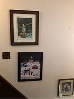Wall display showing two framed pictures: one depicting a rustic scene with a crane and water, and the other a winter village scene in a black frame
