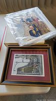 Two items on a table: a framed print and an object wrapped in plastic, likely an unframed print.