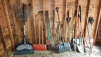 Assorted yard tools in wooden shed showing variety of rakes, shovels, pitchfork, edger and large orange shovel.