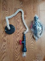 One pendant light laid flat showing the black circular ceiling mount, thick natural rope, black metal pipe body with red valve handle, and clear bulb attached.