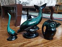 Overall photo of the three Blue Mountain aquatic figurines including the dolphin, puffin creamer, and long necked goose on a wooden floor with furniture background.