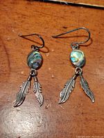 Pair of vintage First Nations sterling silver earrings with feather pendants and oval mother of pearl inlay displayed on wooden surface.
