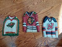 Three ceramic NHL sweaters laid on wooden floor, each with metal hanger and real thread entwined, showing front logos and colors.