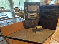 Photo showing two karaoke stereo systems on a table with a large box of CDs and several cassette tapes stacked in front.