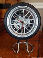 Front view of a vintage-style alarm clock resembling a car tire and alloy wheel, showing clock face with numbers and hands.