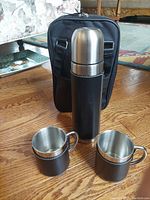 Stainless steel coffee thermos with black leather sleeve and two matching stainless steel cups in leather wraps on wood floor, with black carrying case behind.