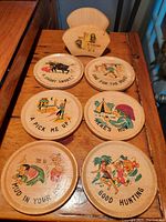 Top view of all six round wooden coasters with colorful cartoon illustrations and sayings arranged on wood surface alongside the matching wooden holder.