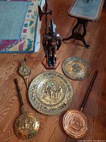 Wide view showing all items in the lot including 2 brass and copper bed warmers, 2 brass plaques, knight poker holder, fireplace bellows, and a brass poker.