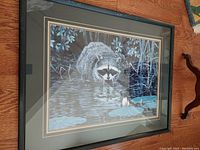 Front view of the framed artwork showing the raccoon in water with lily pads, surrounded by leaves under glass with a wooden frame.