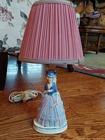 Full view of vintage porcelain figural lamp with attached pink fabric pleated lampshade, showing base figure of a woman in historic dress.