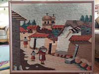 Full view of framed wool textile featuring Mesoamerican village scene with figures and buildings