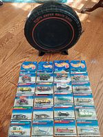 Black tire-shaped Hot Wheels Super Rally Case with red text, displayed behind twenty Hot Wheels cars in original packaging laid out on wooden floor