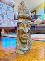 Photo showing vintage hand-carved wood sculpture of a Mesoamerican face with detailed headdress, set on a wooden floor with background furniture.