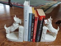 Front view of vintage white porcelain Scottie dog bookends holding six hardcover and paperback novels between them on a wooden floor.