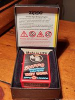 Red Zippo lighter with colorful graphic saying 'Zippo Windproof Lighters They Work Zippo Canada' sitting in original black presentation box with inside lid showing product information and warnings.