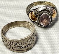 Two silver rings displayed on fabric. One is a wide etched band, the other a gemstone ring with silver and gold detailing.