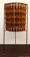 Front view of vintage 1960s teak veneer basket plant stand with brass wire frame on a dark surface against a plain wall.