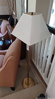 Full view of the brass floor lamp beside a peach upholstered chair, showing the base, shaft and white square tapered lampshade.
