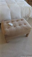 Photo of beige upholstered ottoman placed on carpet near bed, showing full ottoman and legs.