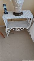 Close-up image of one wicker stand showing top surface and metal scrollwork legs with a lamp and cordless phone on top.