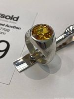 Close-up of the sterling silver ring with yellow faceted stone on a metal clip alongside auction label 149.