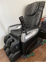 Front-left angle view of the full black massage chair showing seat, backrest, armrests, foot massager and wired remote