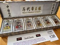 Six hand painted crystal snuff bottles featuring pictures of Chinese monarchs, displayed in a segmented fabric-lined box with informational card and Chinese calligraphy decorations on box interior.