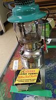 Full view of Coleman camp lantern showing vent top, glass globe, and fuel tank with control knob, alongside boxed Coleman filter funnel.