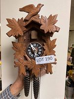 Front view of the wooden cuckoo clock showing bird on top, leaf carvings around clock face, pinecone weights and part of the chain.